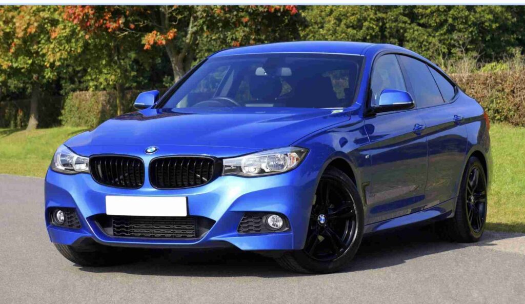 A metallic blue BMW coupe parked on the side of a road with greenery in the background.
